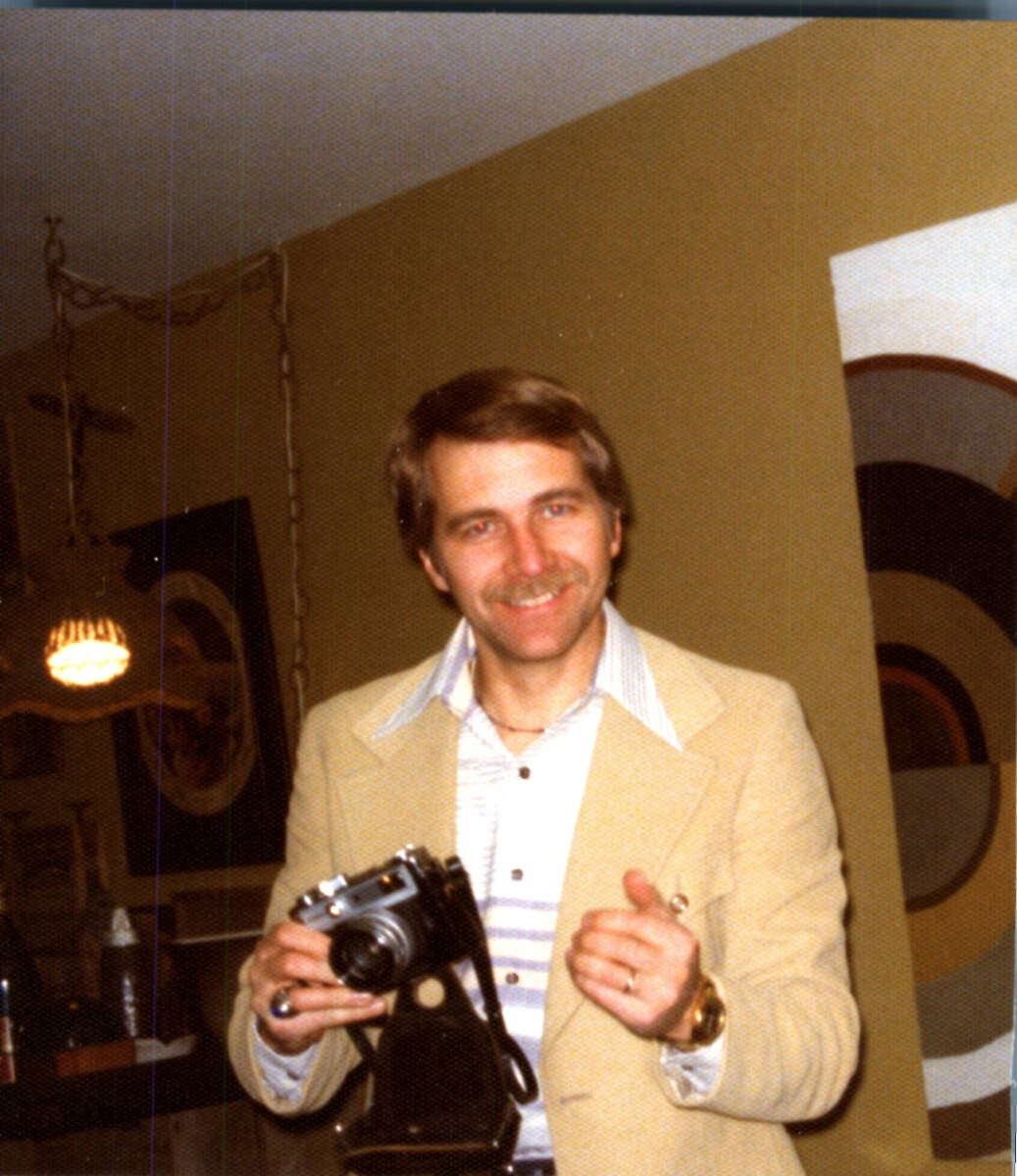 A smiling man in a beige suit holds a vintage camera indoors. The background features modern art on the wall and hanging lights, creating a warm, retro atmosphere.