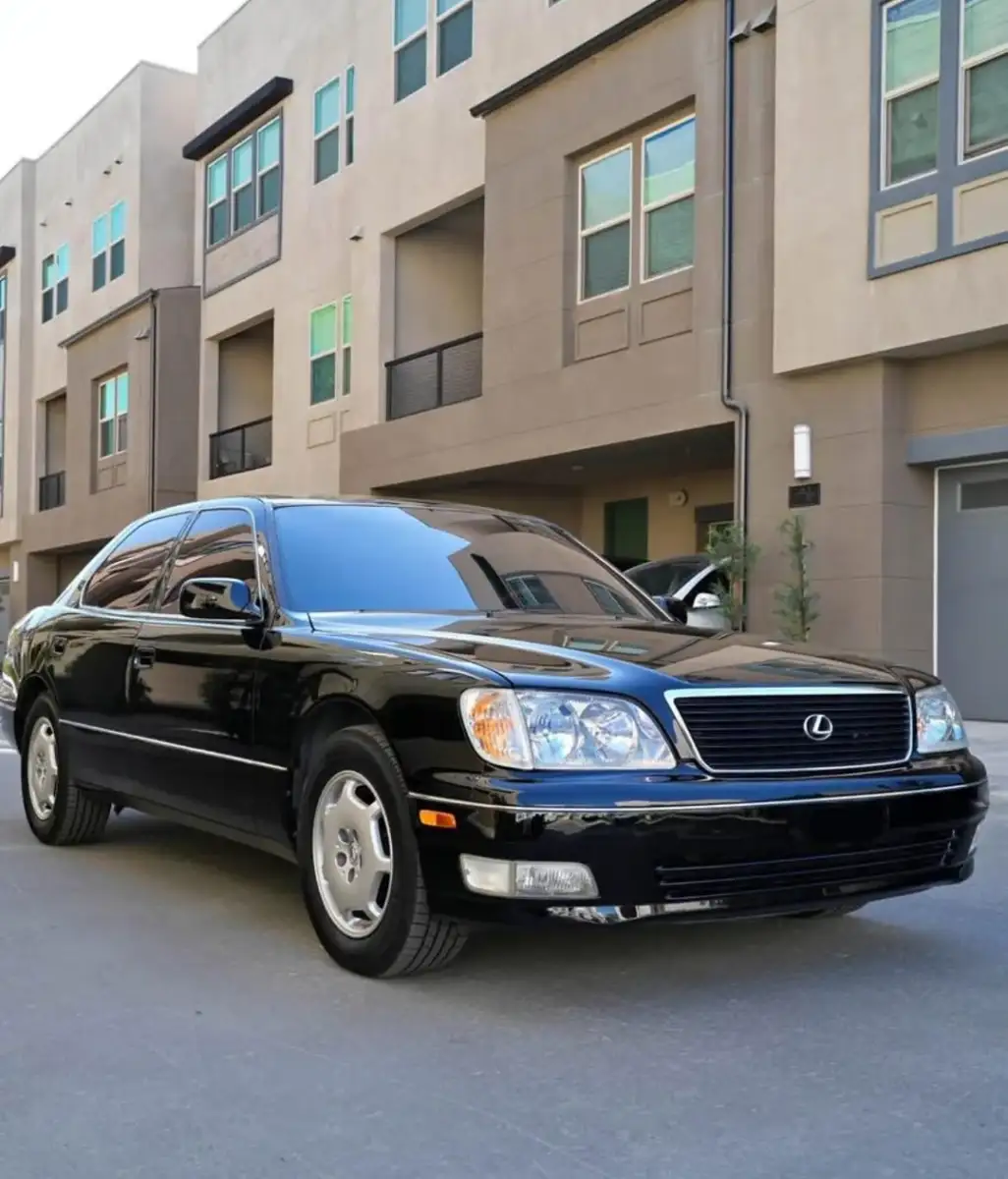 A black Lexus LS sedan is parked on a clean street in front of modern tan apartment buildings with large windows and balconies.