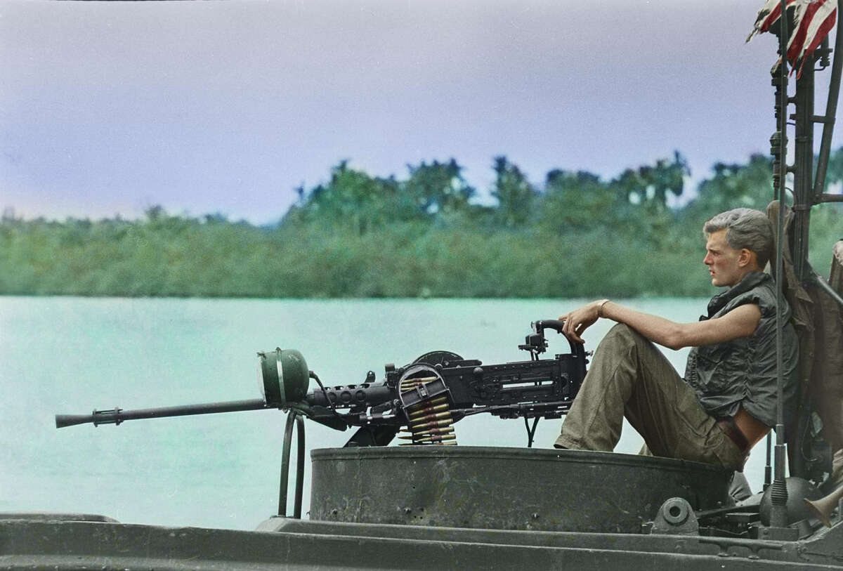 A person in military attire sits on a boat beside a mounted machine gun, gazing toward the water. Lush green trees and an overcast sky are visible in the background.