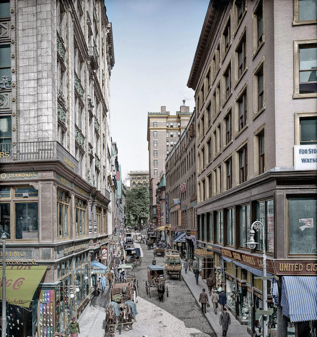 A busy early 20th-century city street scene with people walking, horse-drawn carts, and storefronts with colorful signs lining tall buildings on both sides.