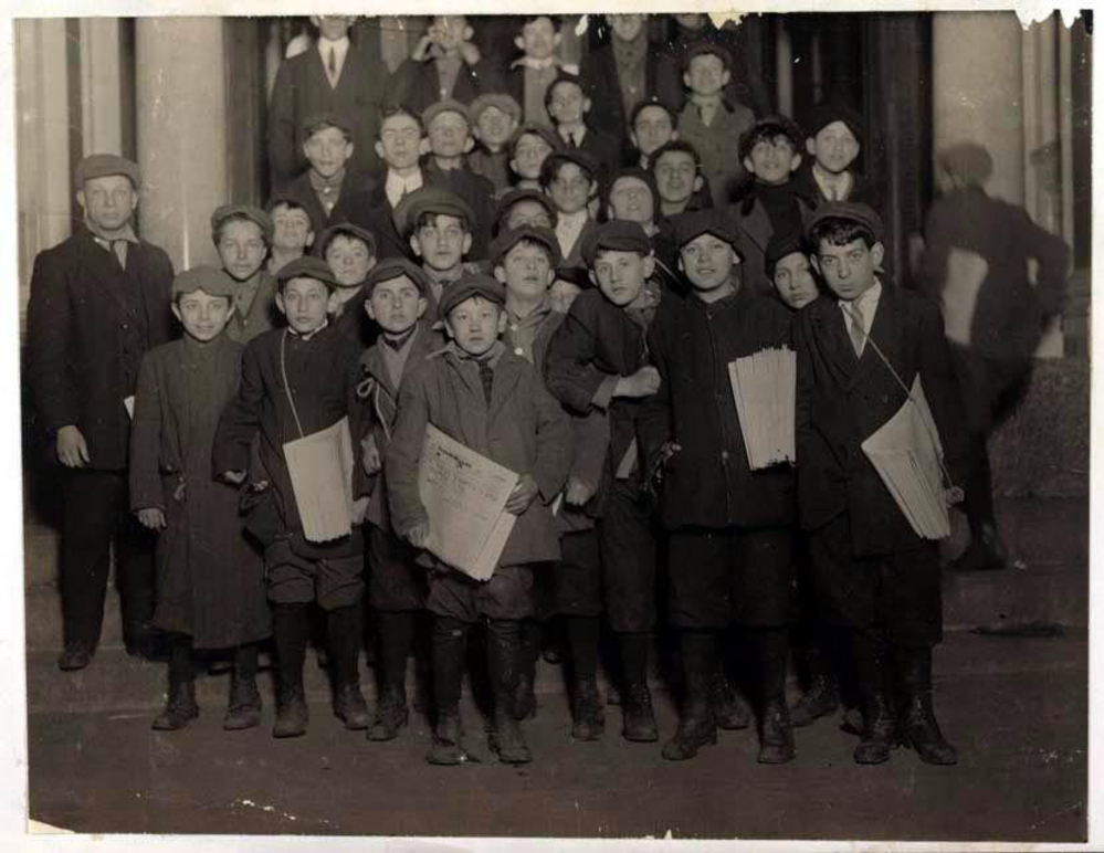 A group of boys, many holding newspapers, stand closely together on a city sidewalk in front of a building, looking toward the camera. Most are wearing hats and coats, suggesting a historical setting.