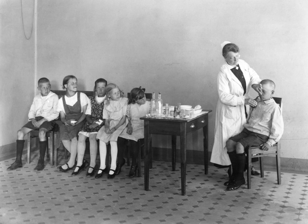 A nurse in a white uniform administers medicine to a seated boy, while five other children sit in a row watching, in a sparsely furnished room with a tiled floor and a table holding medical supplies.