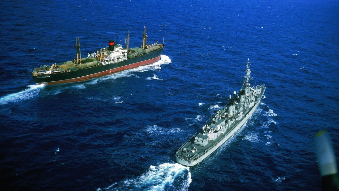Aerial view of a military ship and a cargo vessel traveling on deep blue ocean waters, creating white wakes behind them under clear skies.