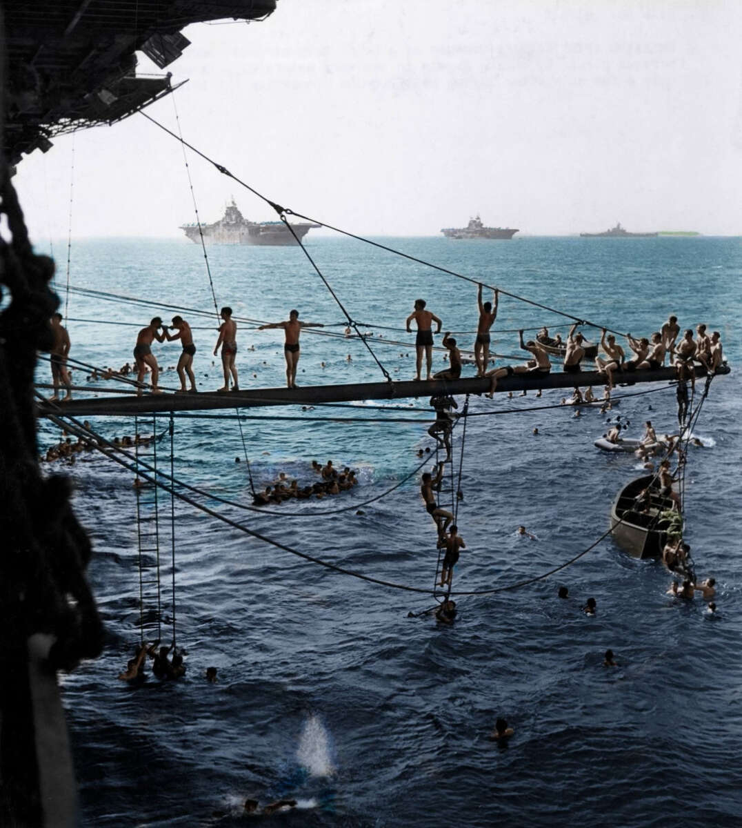 Sailors in swimsuits dive, swim, and climb ropes and ladders beside a large ship at sea. Several ships are visible on the horizon under a clear sky. Some sailors are on a wooden platform above the water.