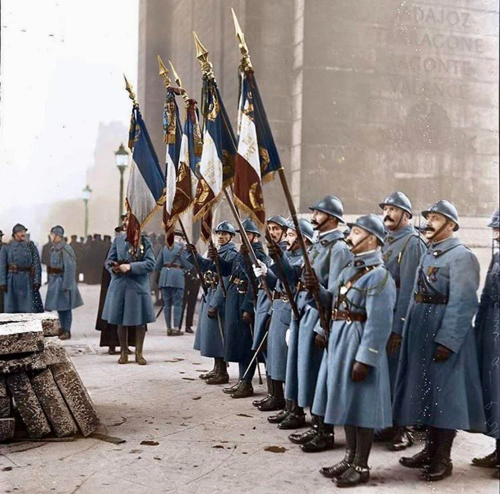 A group of World War I-era French soldiers in blue uniforms and helmets stand in formation, some holding large, colorful flags. They appear to be participating in a ceremony near a stone monument.