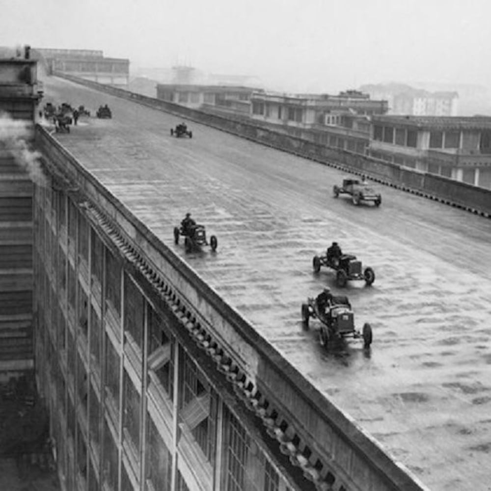 Vintage cars drive on a wide, sloped rooftop track atop a large industrial building, with steam rising on the left and city structures visible in the background under a cloudy sky.