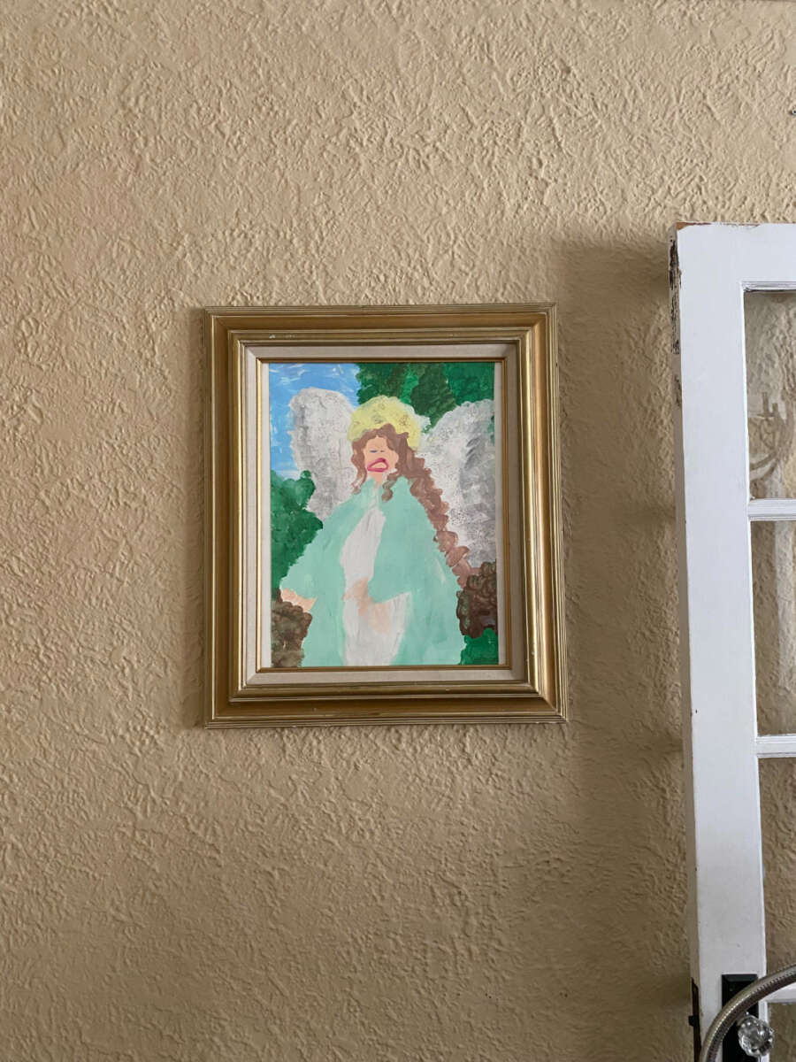A framed painting of an angel with green wings, a yellow halo, and long brown hair hangs on a textured beige wall next to a white door with glass panes.