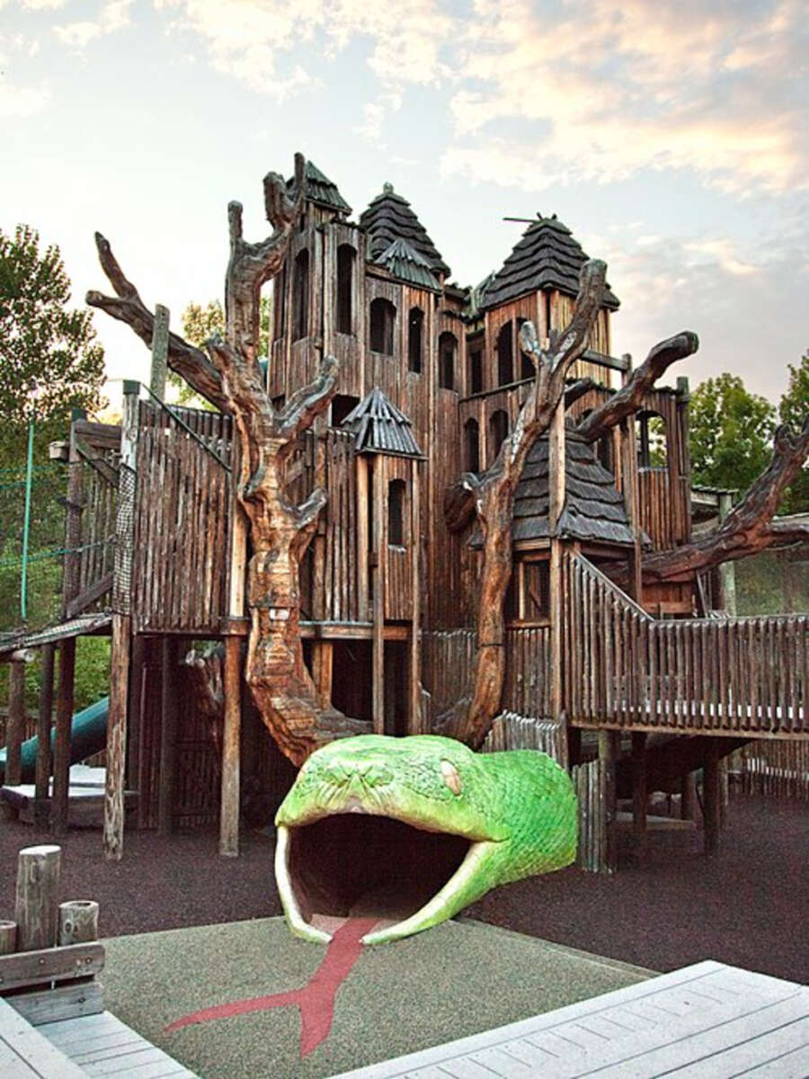 A large wooden playground structure shaped like a fantasy castle with twisted branches and a slide resembling a giant green snake with its mouth open and red tongue extended. The sky is partly cloudy at sunset.