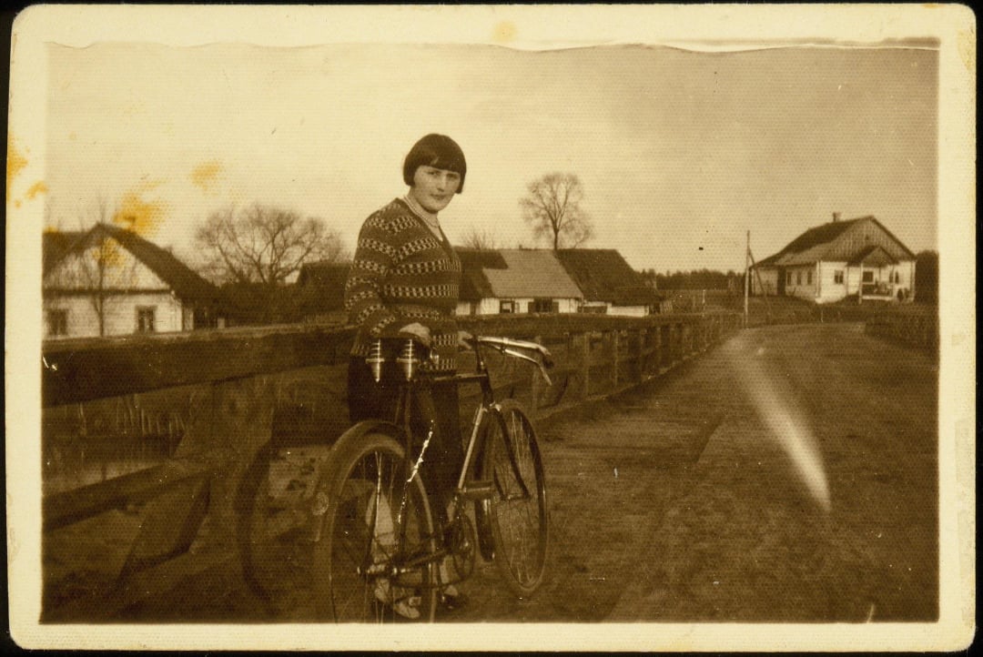 A person with short hair and a patterned sweater stands holding a bicycle on a wooden bridge in a rural village, with houses and leafless trees in the background, in a sepia-toned vintage photo.