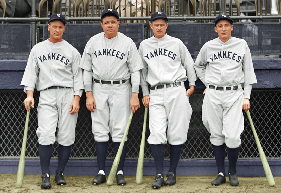 Four New York Yankees baseball players in classic uniforms stand side by side, each holding a bat, posing for a photo at a stadium.