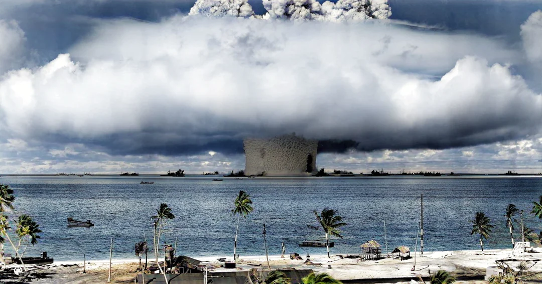 A large mushroom cloud rises over the ocean at a distance, with palm trees and beach huts in the foreground and several boats on the water under a mostly cloudy sky.