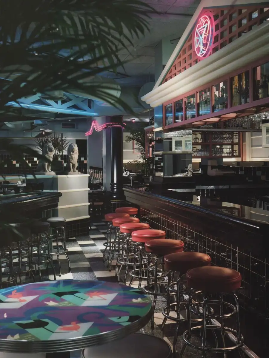 A retro-style bar with red round stools along a dark tiled counter, neon lights, checkered floor, decorative statues, and a colorful abstract table in the foreground. The atmosphere is vibrant and nostalgic.