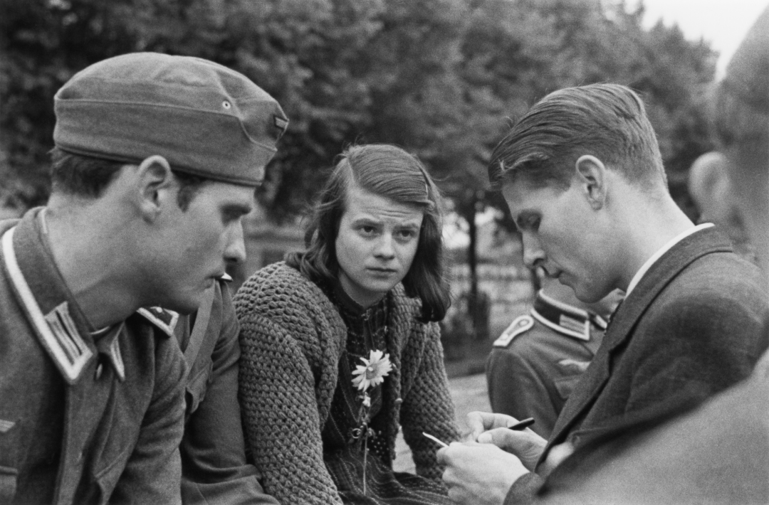 A young woman sits between two young men in military uniforms outdoors. The woman, wearing a crocheted sweater and a flower pin, looks directly at the camera while the men appear focused on writing or talking. Trees are visible in the background.