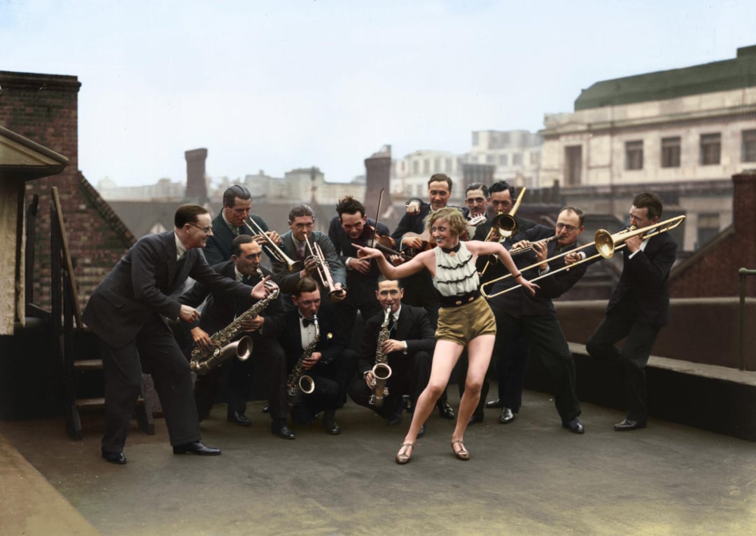 A woman in a striped top and shorts energetically dances on a rooftop, surrounded by a lively jazz band of men playing brass and woodwind instruments. Buildings are visible in the background.