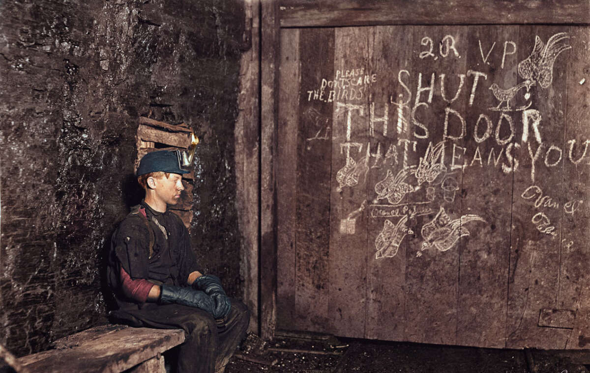 A coal miner wearing a helmet and gloves sits on a wooden bench in a dim, dirty underground tunnel. Behind him, a wooden door is covered with chalk messages and bird drawings, including "SHUT THIS DOOR THAT MEANS YOU.