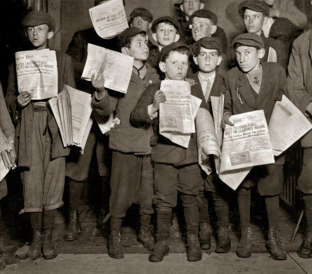 A group of boys dressed in early 20th-century clothing stand together, holding newspapers, some with papers under their arms and others waving them, looking in different directions.