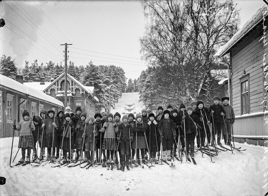 A group of children dressed in winter clothing stand in the snow on a street between wooden houses, holding skis and ski poles, with trees and snowy rooftops in the background.