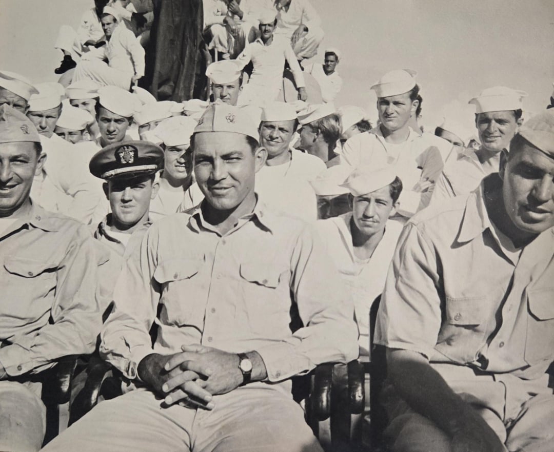 A group of uniformed sailors and one officer sit and stand closely together outdoors, smiling and looking toward the camera under a bright sky.