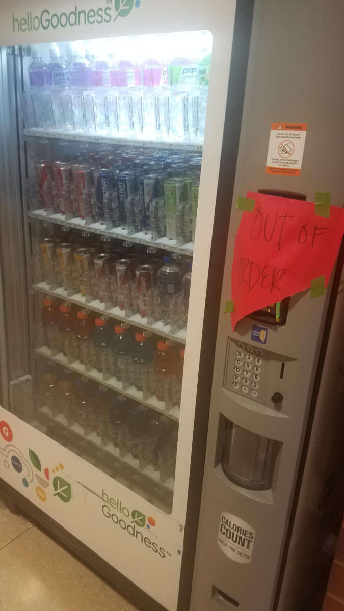 A vending machine filled with bottled drinks has a bright red "OUT OF ORDER" sign taped near the keypad, indicating it is not working.