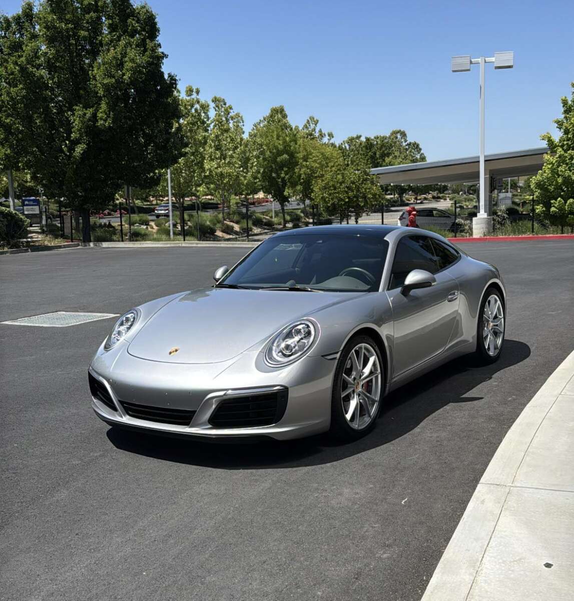 A silver Porsche sports car is parked on a sunlit asphalt road near a curb, with green trees and a covered walkway visible in the background.