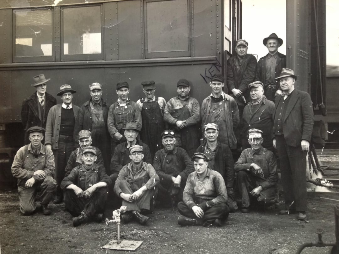 A black-and-white photo of 18 men in work clothes and caps posing in front of a train car. Some are standing, others kneel or sit on the ground. The group appears to be railroad or train workers from an earlier era.
