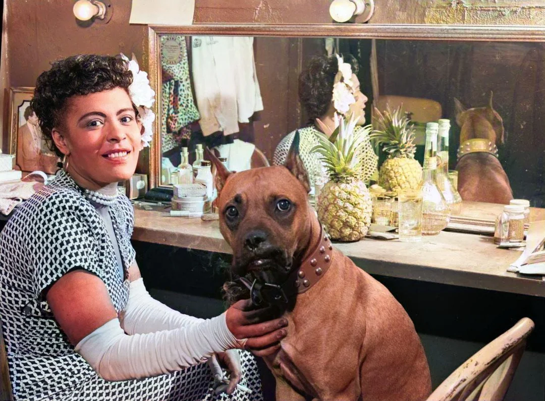 A woman in a checkered dress with white gloves sits at a vanity table, smiling, while a brown dog with a studded collar sits beside her. The table holds bottles, a pineapple, and a mirror reflecting the scene.