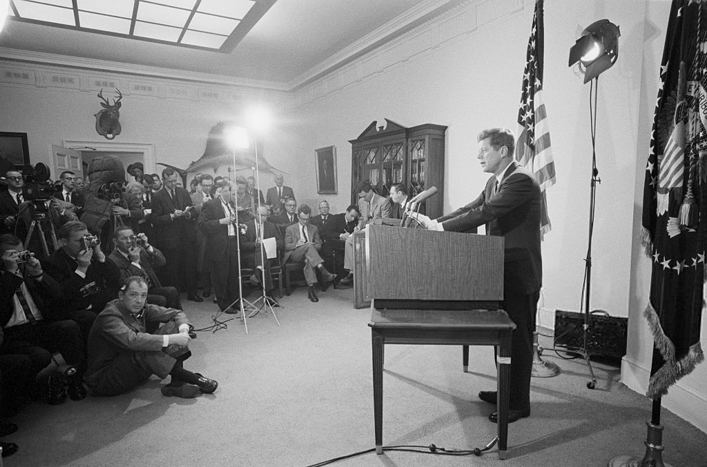 A man speaks at a podium in a formal room with an American flag and press gathered around, including photographers, reporters, and cameramen capturing the event. Bright studio lights illuminate the scene.