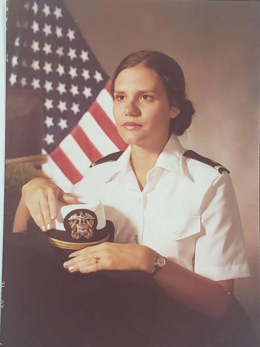 A woman in a white military uniform sits beside a table, holding a hat with an insignia. An American flag is in the background. She has brown hair styled in a bun and wears a watch and a ring.