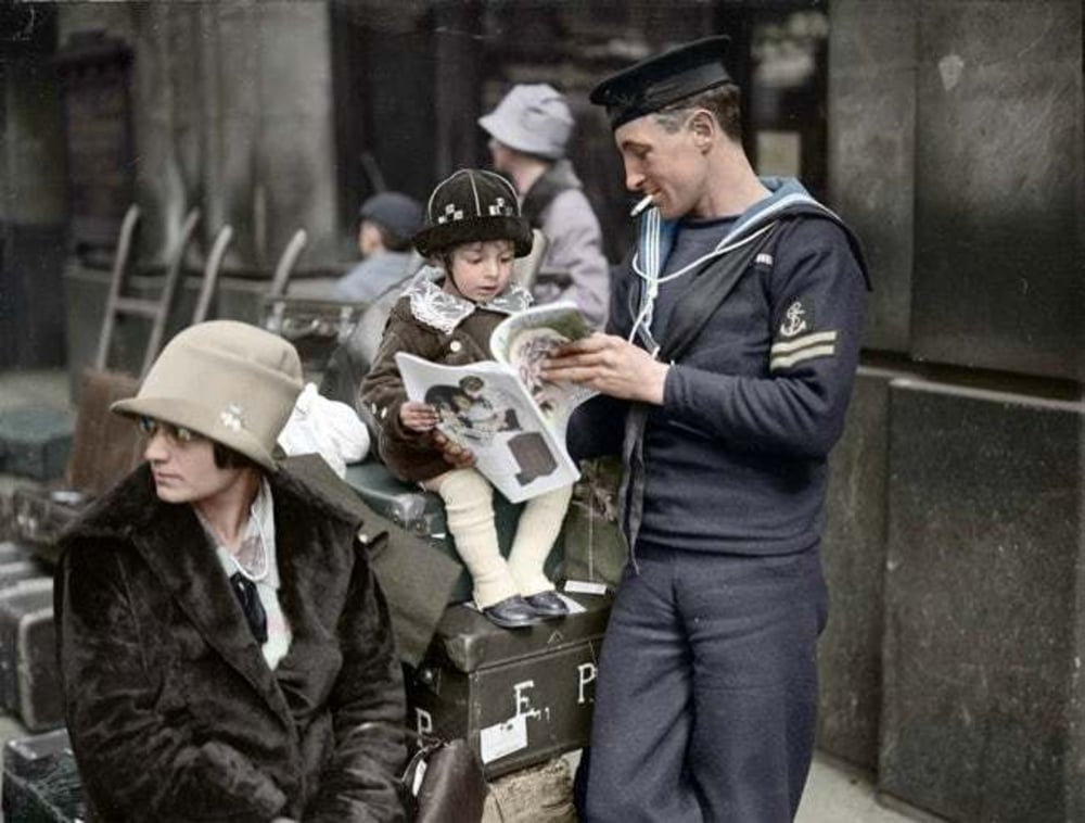 A sailor in uniform reads a magazine while standing beside a woman and a child sitting on luggage outside. The woman wears a cloche hat and coat; the child also has a hat and looks at the sailor. Other people are blurred in the background.