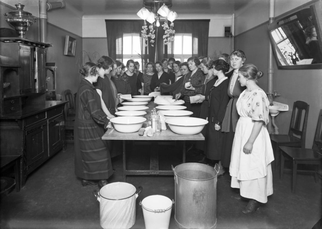 A group of women gathers around a long table set with large white bowls and jars in a classroom or kitchen setting, preparing for a lesson or demonstration. The room is simply furnished with chairs and cabinets.