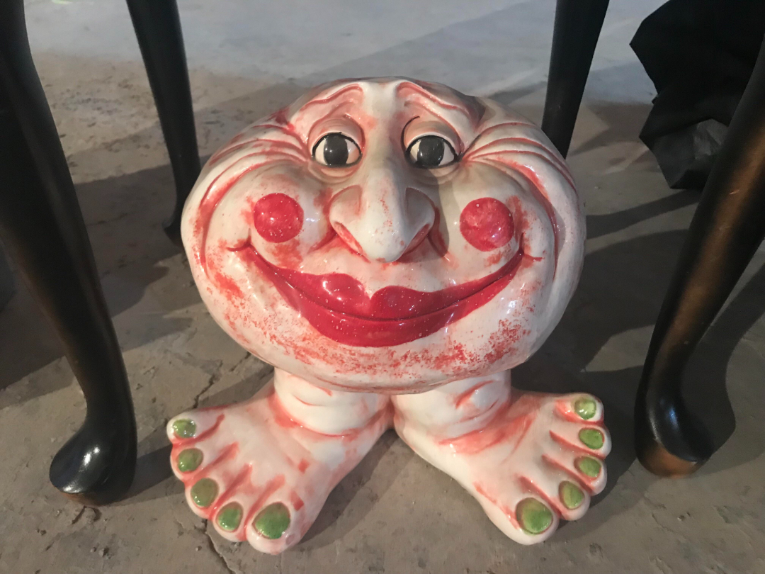 A whimsical ceramic sculpture with a round, smiling face, rosy cheeks, red lips, wide eyes, and two feet with green toenails, placed on a concrete floor between dark wooden chair legs.