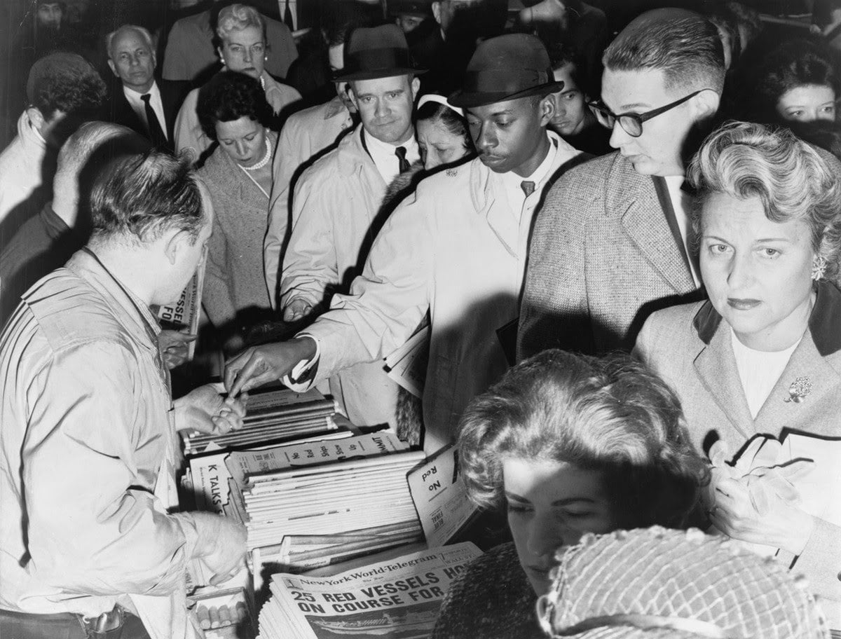 A crowd of people, mostly in coats and hats, gathers around a newsstand, eagerly buying newspapers. The image is in black and white and appears to be from the mid-20th century.