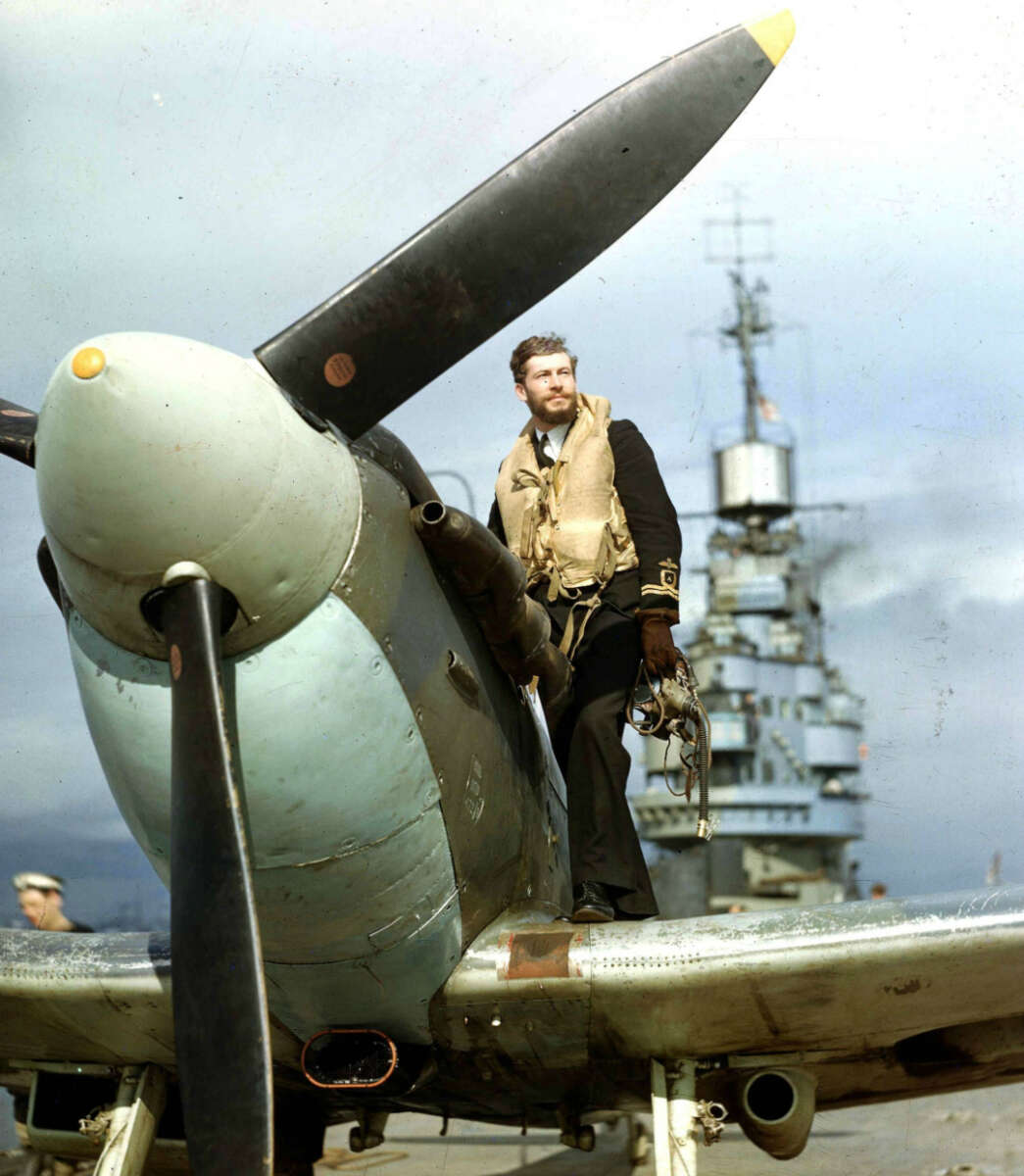A man in a military uniform and life vest stands on the wing of a propeller aircraft, holding a helmet, with a large naval ship in the background under a cloudy sky.
