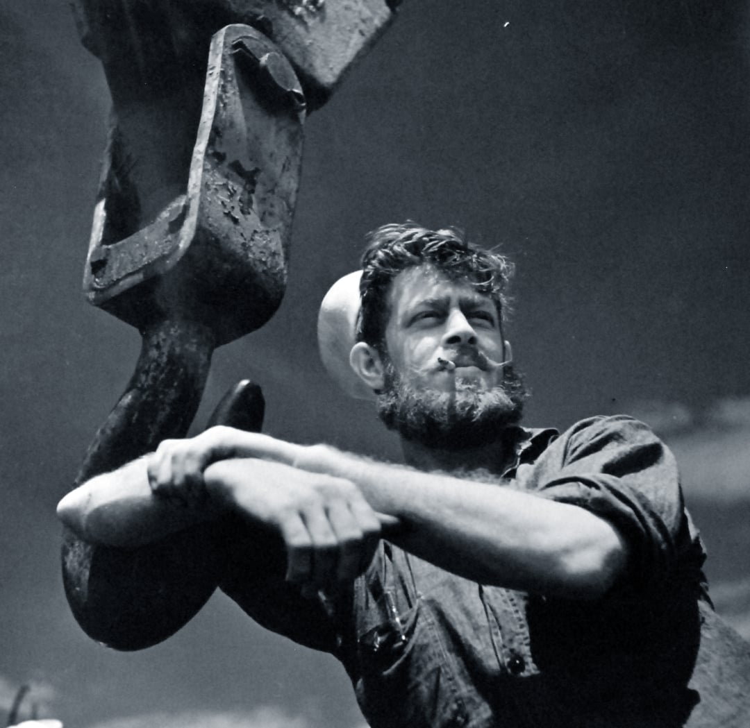 A bearded man in work clothes and a hard hat leans against a large industrial hook, looking off into the distance with a serious expression, set against a slightly cloudy sky.