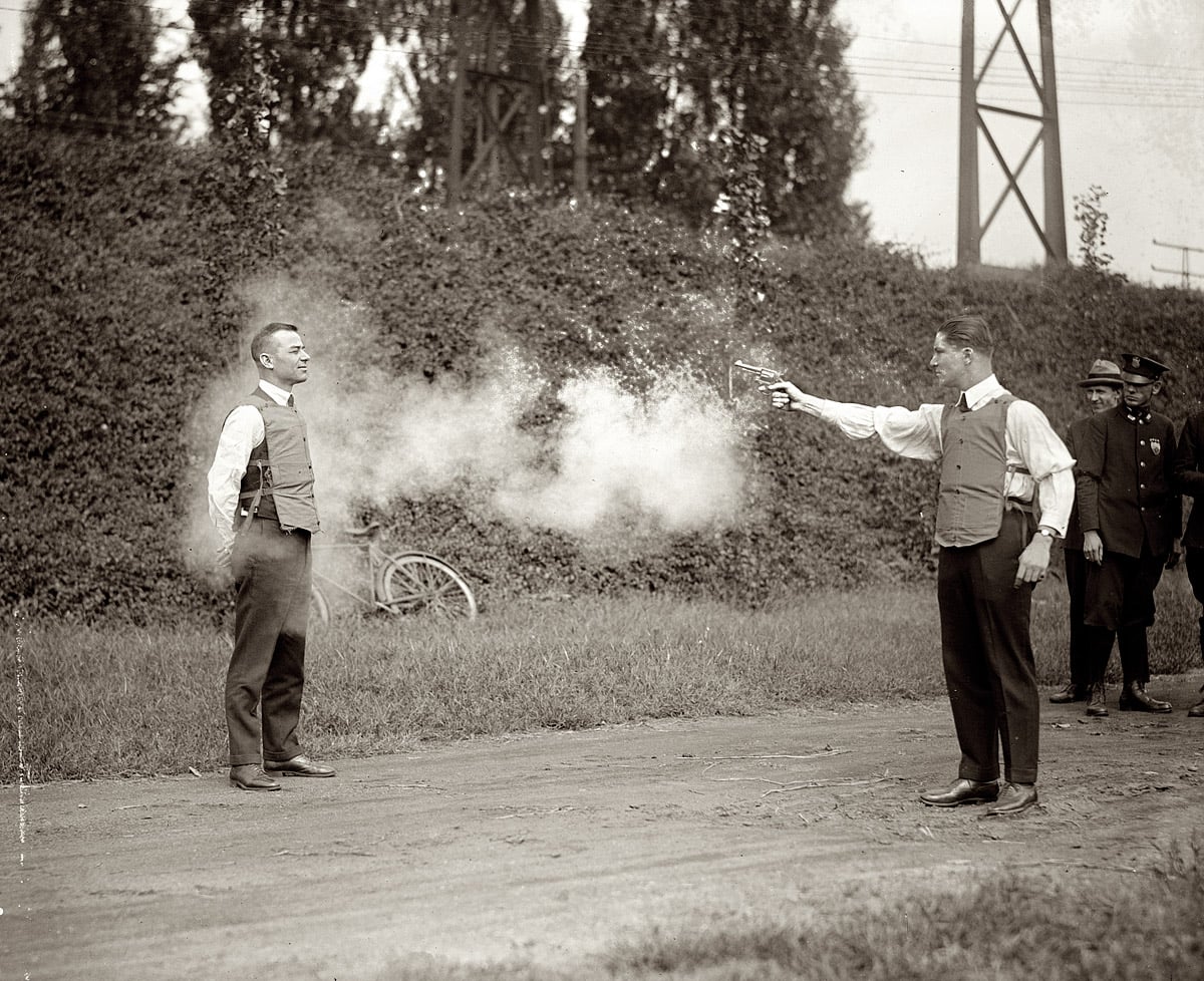 A man fires a pistol at another man's chest at close range outdoors; both wear protective vests while onlookers stand in the background, observing the demonstration.