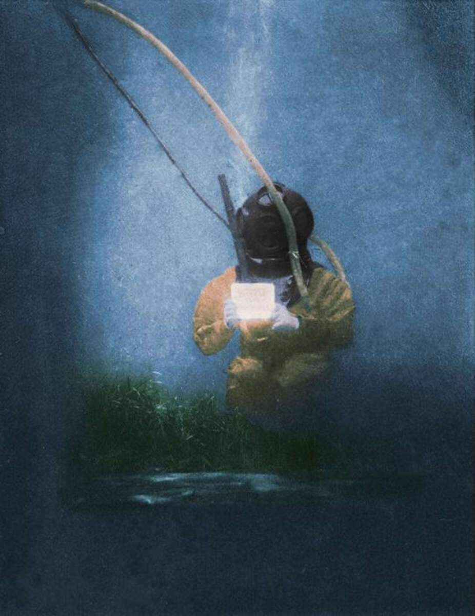 A deep-sea diver in a yellow suit and helmet holds a glowing device underwater, surrounded by blue water with shafts of light and some green plants on the seabed.