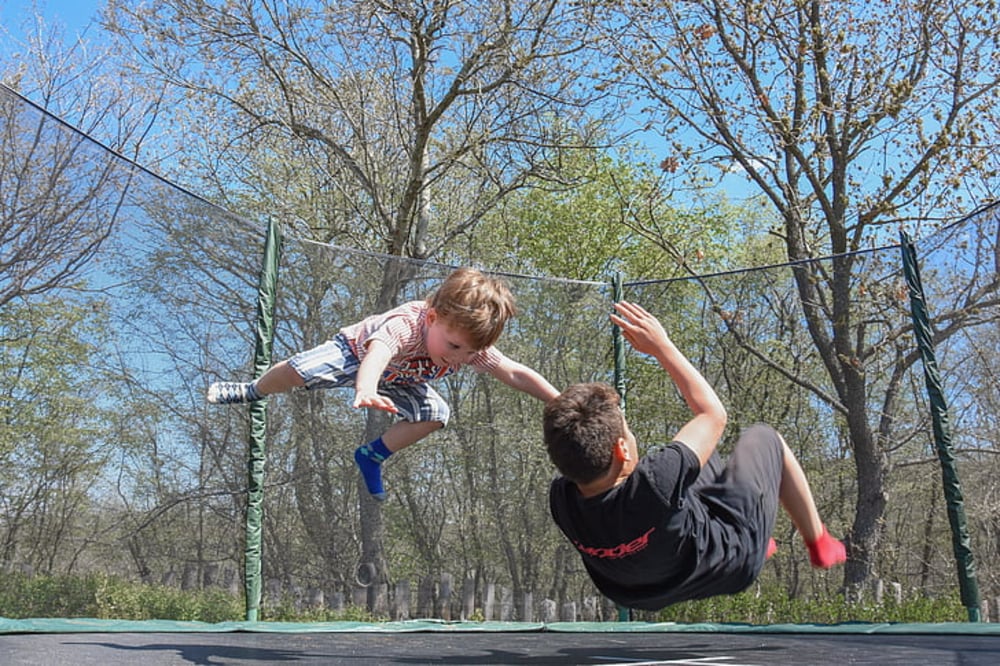 Two boys are mid-air on a trampoline in a backyard, with trees and a blue sky in the background. One boy is flying horizontally while the other is falling backward.