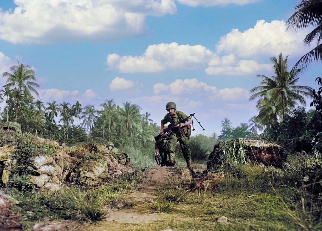 A soldier in green military gear and helmet walks through a tropical landscape with palm trees, holding a rifle. The sky is blue with scattered clouds, and the area appears to be a combat zone with dirt paths and sandbags.