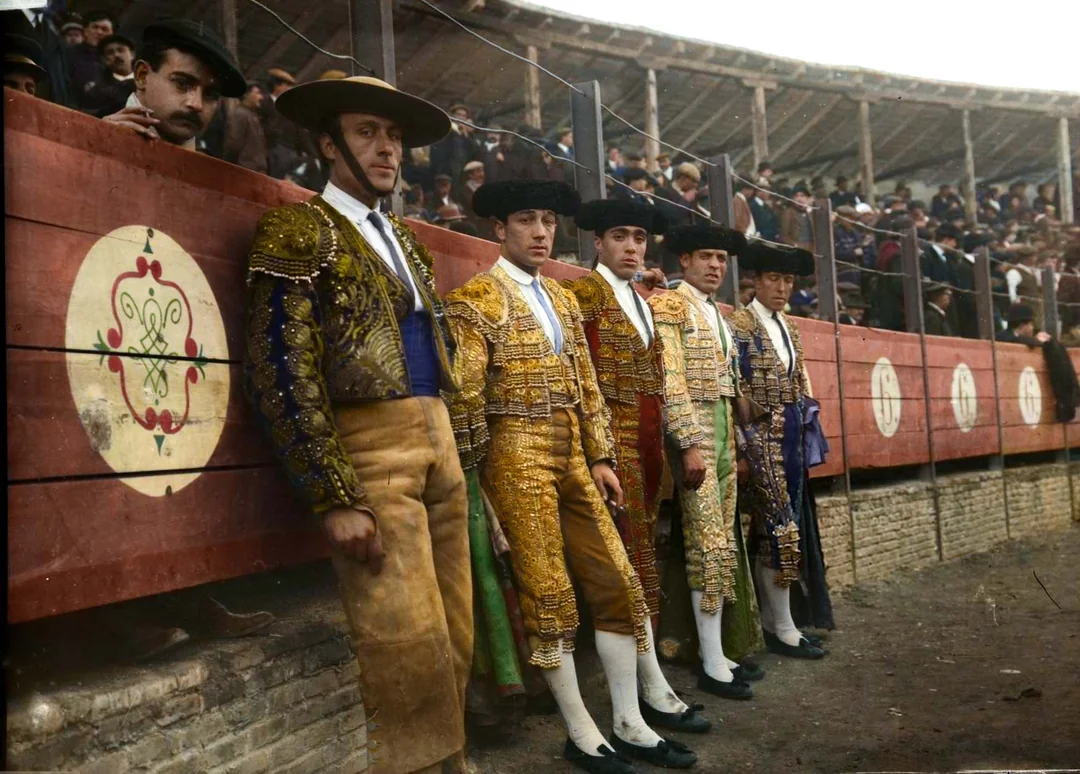 Five bullfighters in ornate traditional costumes stand against a wooden barrier in a bullfighting arena, with a crowd watching from the stands behind them. The scene appears historic and festive.