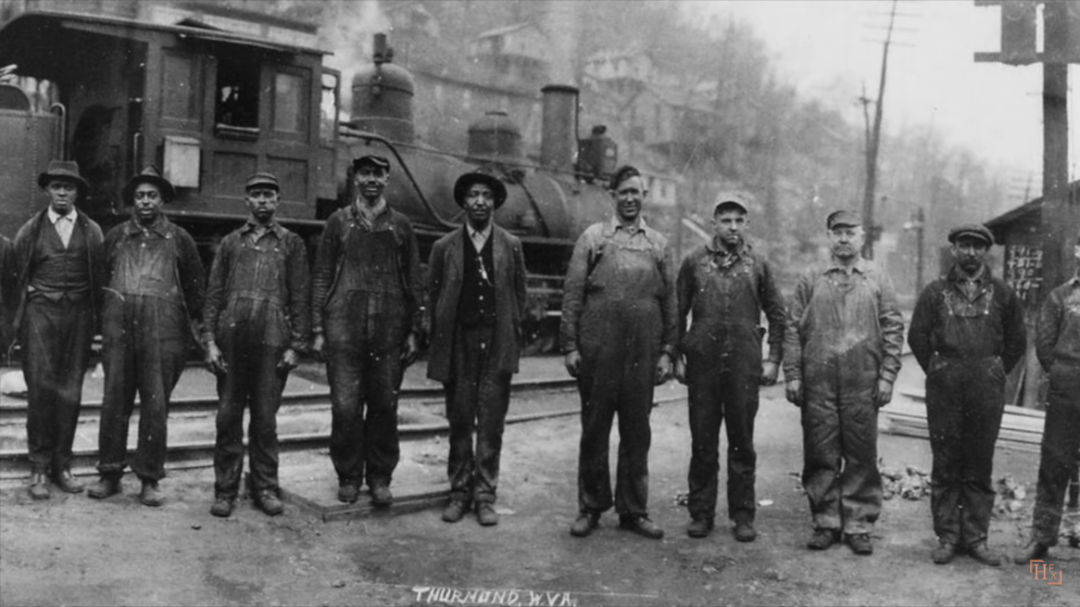 A black and white photo of nine men in work clothing posing on a railway track in front of a steam locomotive in Thurmond, West Virginia. Trees and buildings appear in the background.