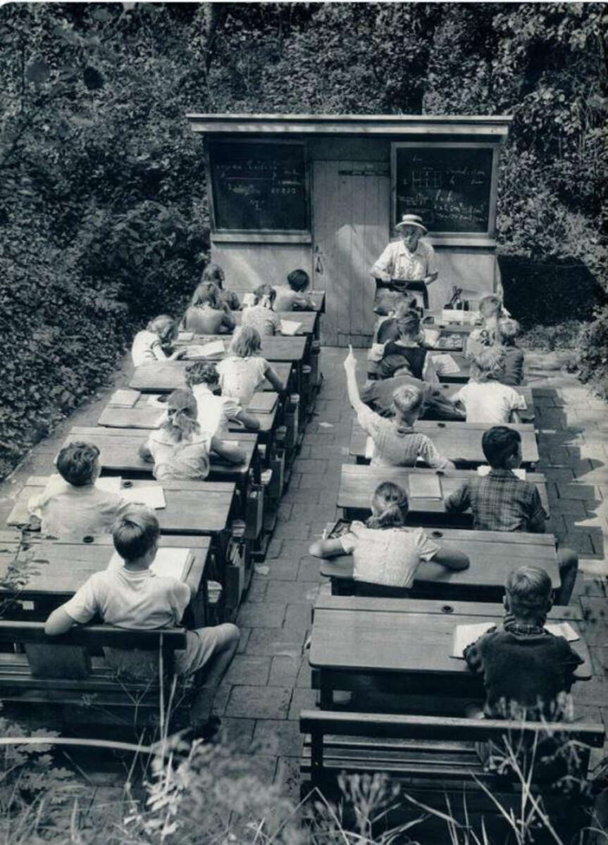 Black-and-white photo of a classroom outdoors, with children seated at desks and a teacher standing by two chalkboards. The class is surrounded by trees; one student raises a hand to answer, and others are writing or listening.