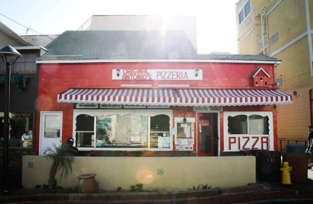 A small, red pizzeria with a striped red and white awning, large front windows, and a sign reading “Original Antonio’s Pizzeria.” There are plants and a trash can outside, and sunlight shines from behind the building.
