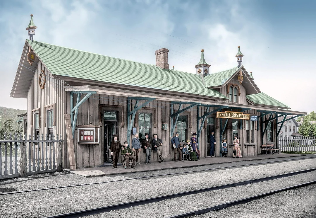 A vintage train station with a green roof and wooden exterior. Several people, including men, women, and children, are waiting on the platform near the tracks under a partly cloudy sky.