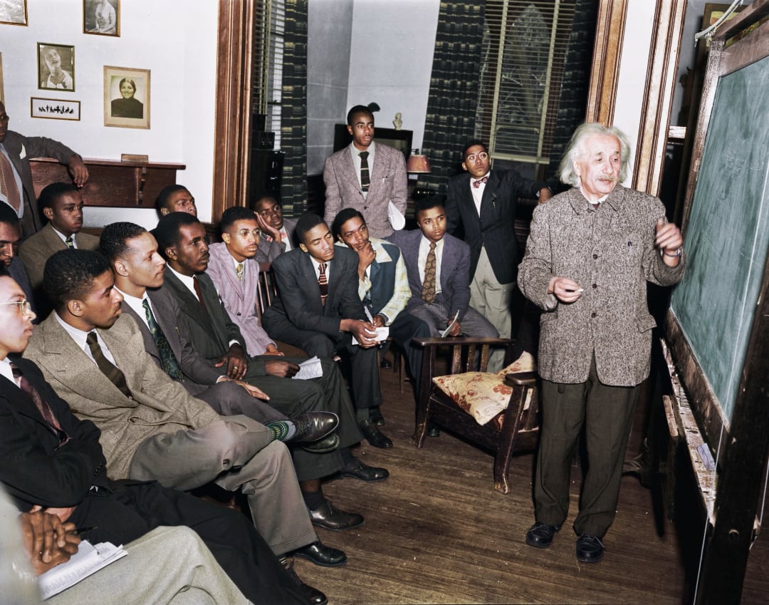 Albert Einstein stands at a chalkboard lecturing to a group of seated young Black men in suits, who are attentively listening and taking notes in a classroom setting.