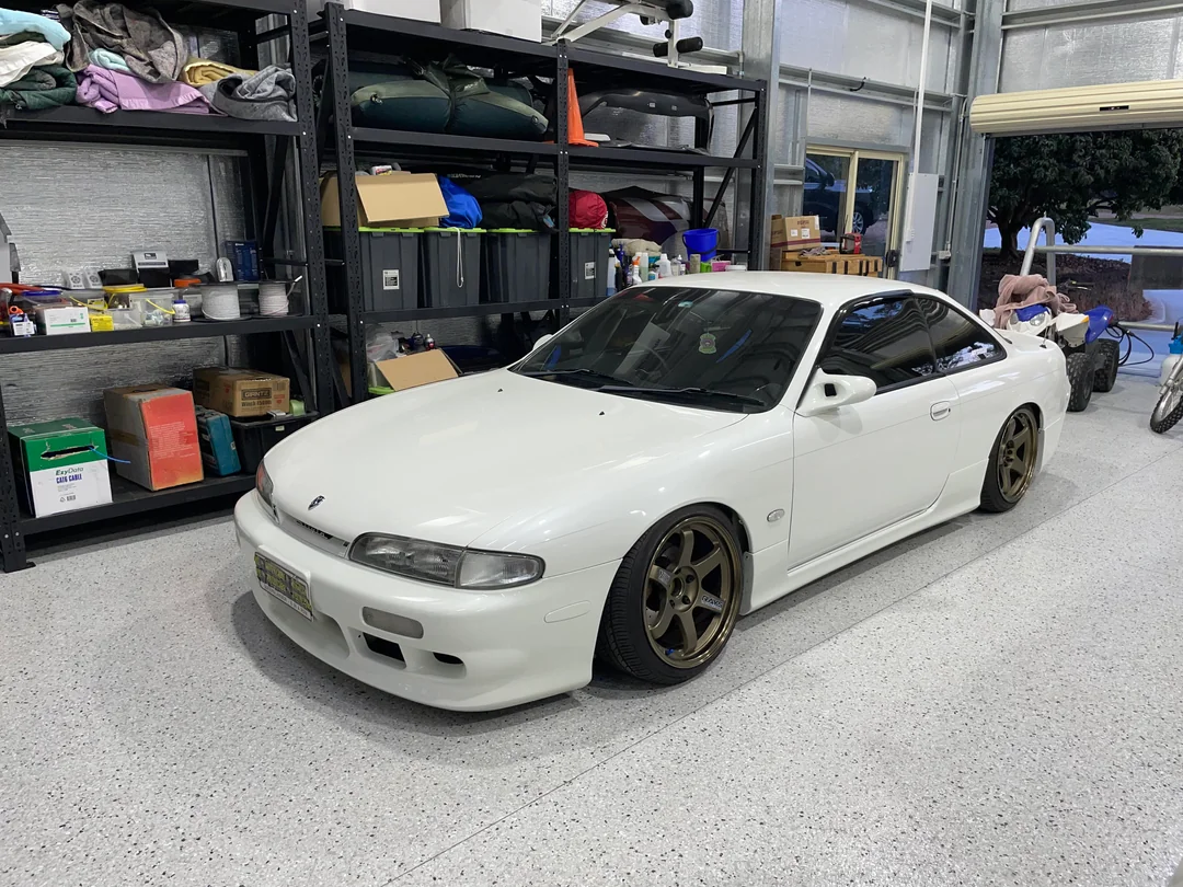 A white Nissan Silvia S14 sports car with bronze wheels is parked in a clean garage. The garage has shelves filled with boxes, tools, and various items. The car has tinted windows and a lowered stance.