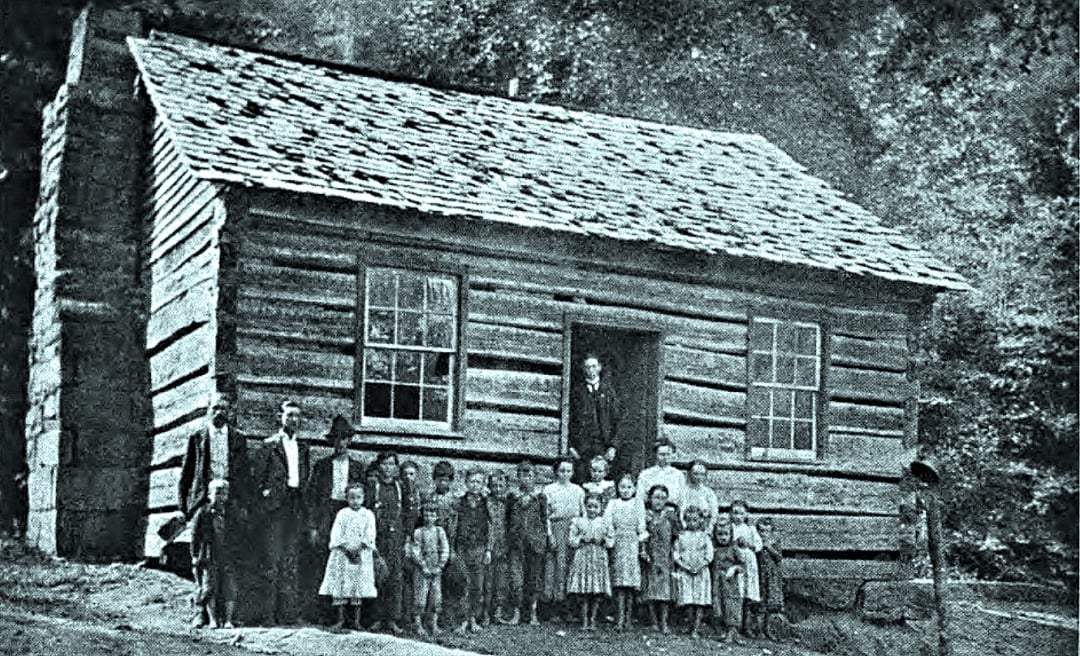 A group of children and adults stand outside a small, old-fashioned log cabin schoolhouse with two windows and a wooden shingles roof, surrounded by trees.
