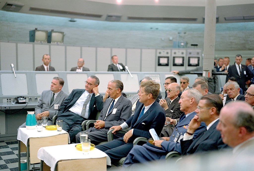 A group of men in suits sit attentively in a control room with monitors and panels in the background. Some have drinks on small tables in front of them, and all appear focused on a presentation out of the frame.