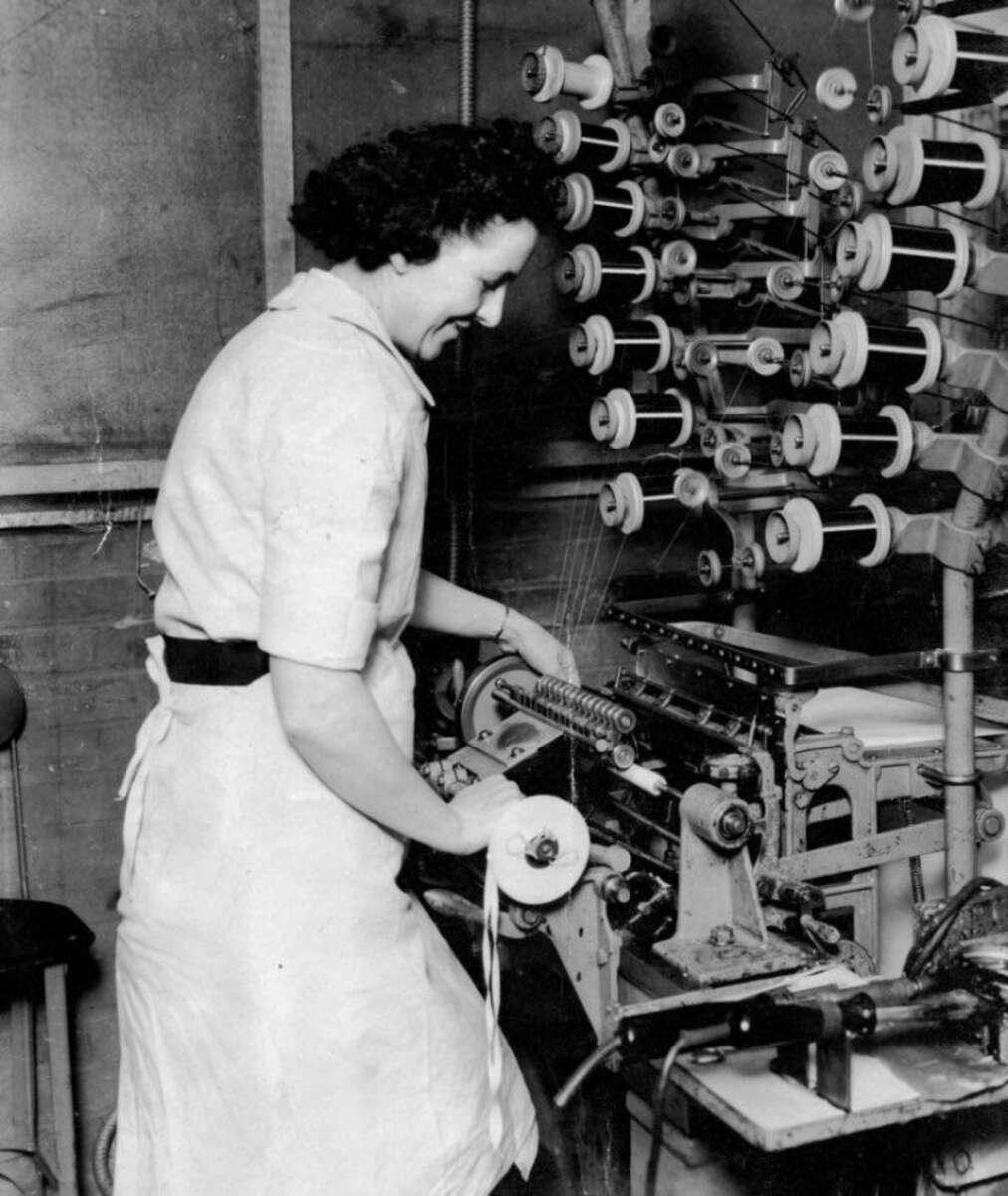 A woman in a work dress operates a textile machine, managing spools of thread in a factory setting. She is smiling and holding a spool while standing in front of machinery with many thread spools.