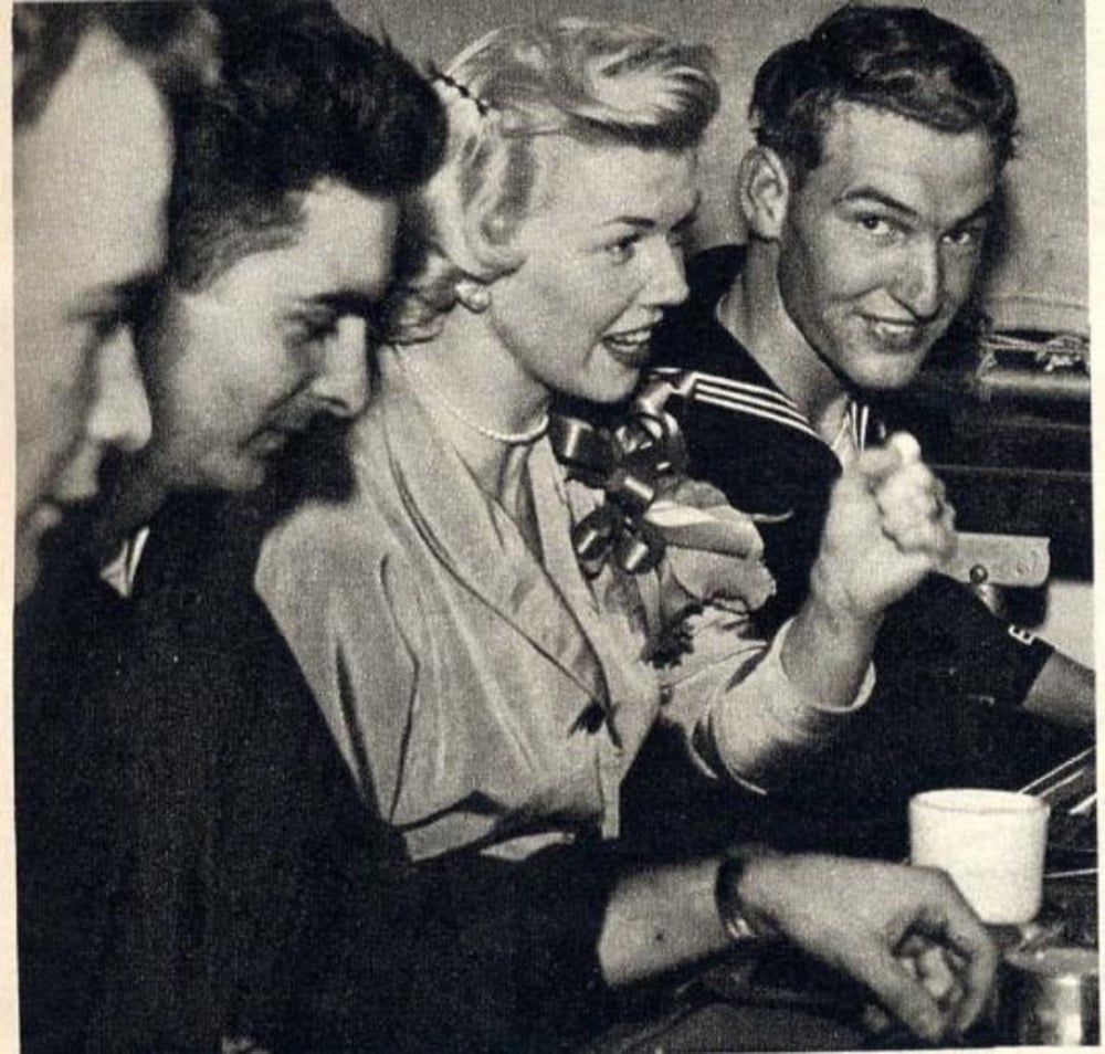 Four people sit closely together at a counter, three men and one woman. The woman, wearing a light jacket and a flower corsage, is talking animatedly while the man in a sailor uniform next to her smiles at the camera.