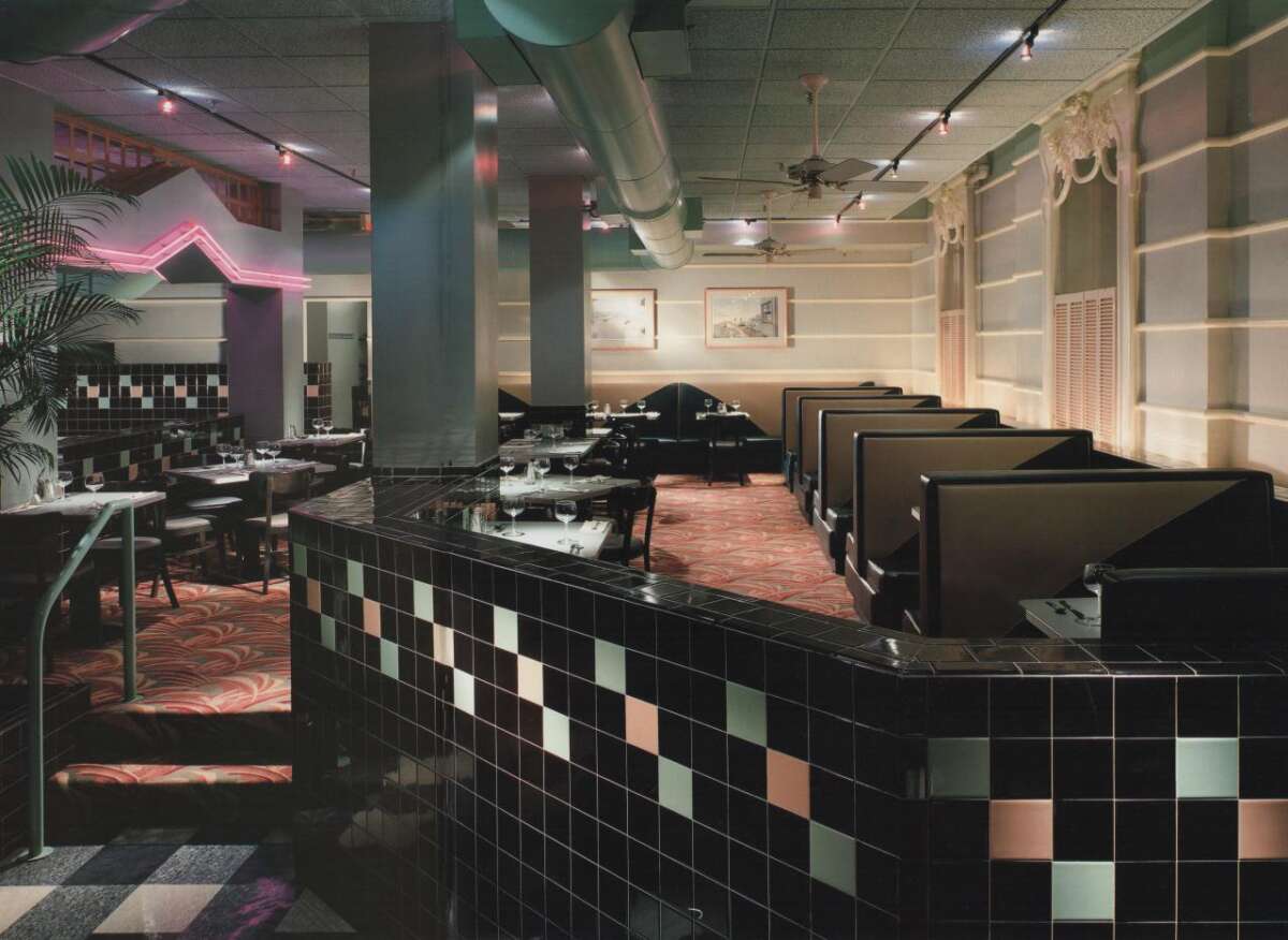 Retro-style diner interior featuring black and pink tiled walls, black leather booths, patterned carpet, and soft neon lighting; tables are set for dining and framed art is on the walls.